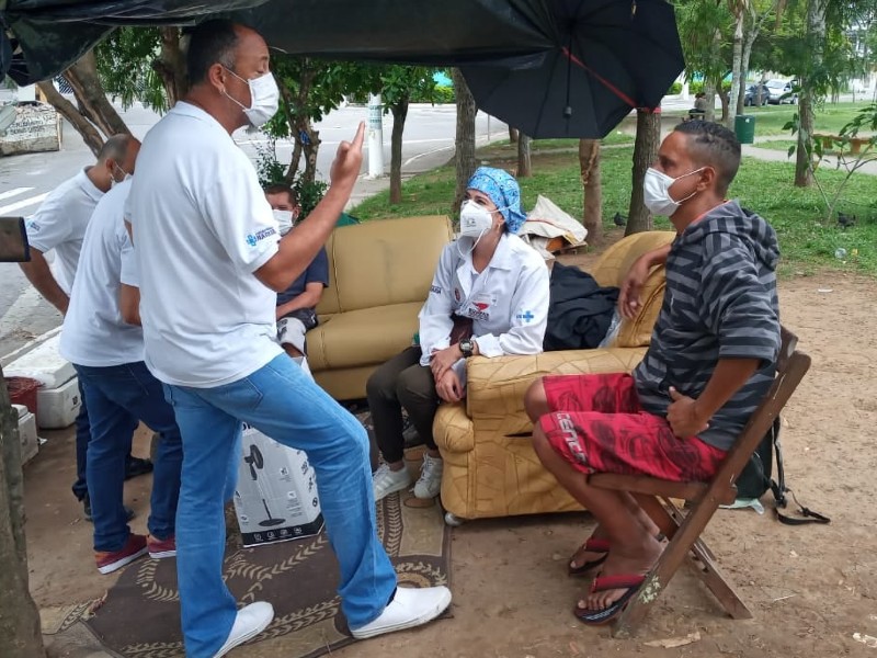 Agente do Consultório na Rua vestindo calça jeans,  camiseta branca e máscara conversa com um morador de rua, que está sentado em sua frente e veste uma bermuda vermelha e um moletom listrado de cinza e preto. Uma agente com jaleco branco, máscara e toca azul acompanha a conversa. Ao fundo, outros dois agentes conversam com um homem sentado num sofá bege